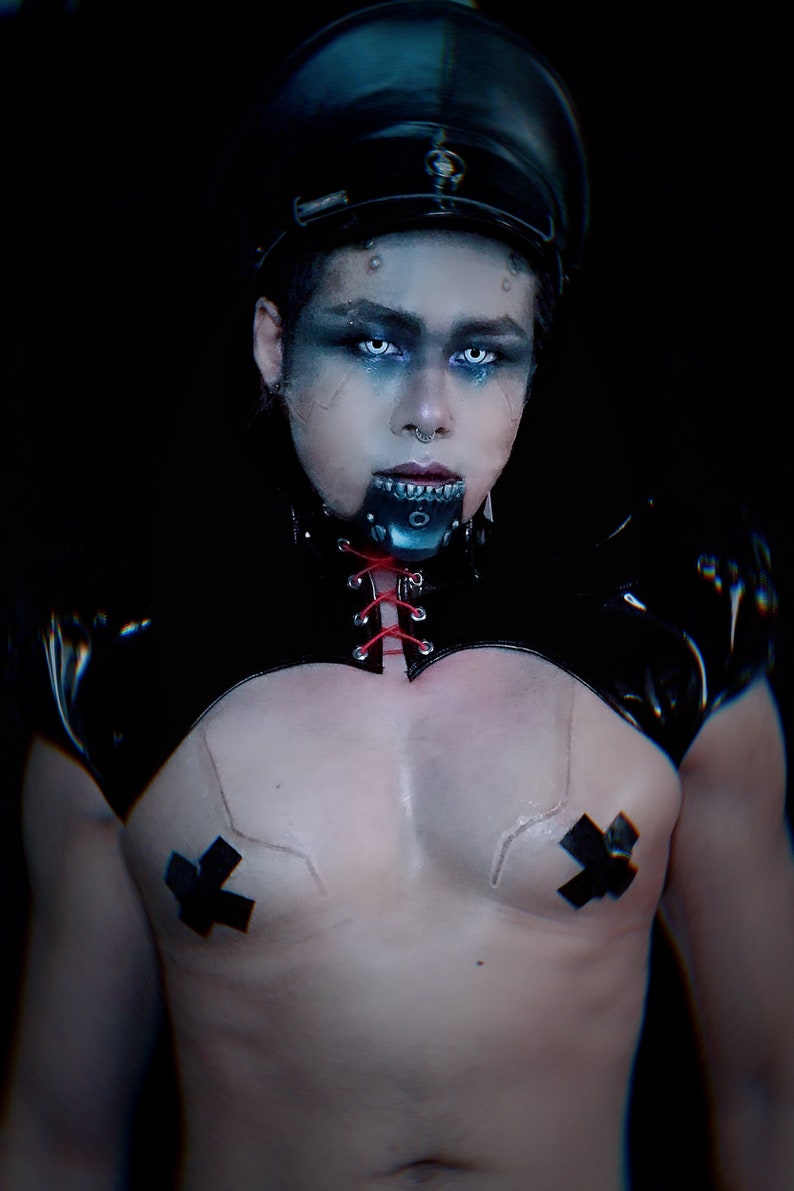 May include: A person wearing a black leather choker with red laces, a black leather top with a lace-up front, and a black cap. They have blue eyeshadow and white contact lenses. They are wearing black tape over their chest.