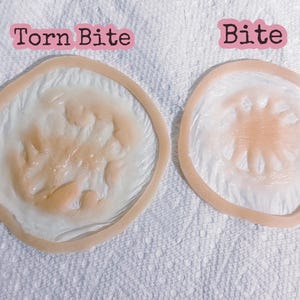 Puede incluir: Dos prótesis de heridas. Una está etiquetada como "Torn Bite" y la otra como "Bite". Ambas son circulares con un borde marrón claro y un interior blanco y color carne, diseñadas para simular lesiones cutáneas.