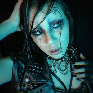 May include: A woman with dark hair and blue-toned makeup wears a black leather jacket with silver studs and a silver chain necklace with large rings. She is wearing a black bra with straps and has black nail polish.