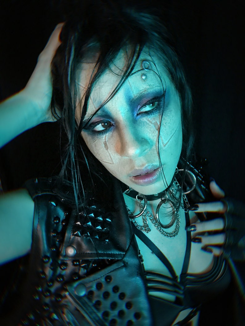 May include: A woman with dark hair and blue-toned makeup wears a black leather jacket with silver studs and a silver chain necklace with large rings. She is wearing a black bra with straps and has black nail polish.