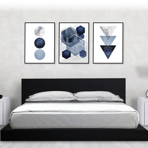 May include: Three framed abstract geometric prints in shades of blue and silver. The prints are hung on a white wall above a black platform bed with gray bedding.