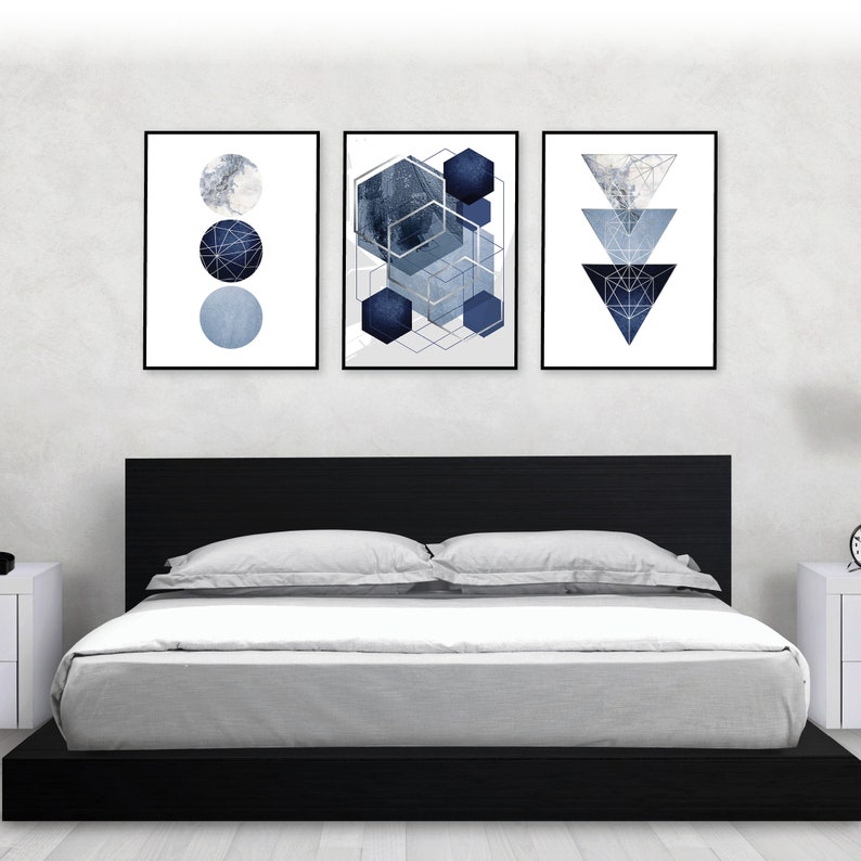 May include: Three framed abstract geometric prints in shades of blue and silver. The prints are hung on a white wall above a black platform bed with gray bedding.