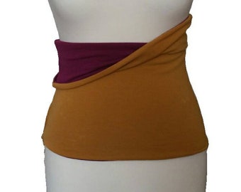 May include: A wide, wrap-style fabric waist warmer or belly band featuring a mustard yellow outer layer and a deep plum purple inner lining. The garment is shown on a white mannequin torso, displaying a layered, overlapping design.