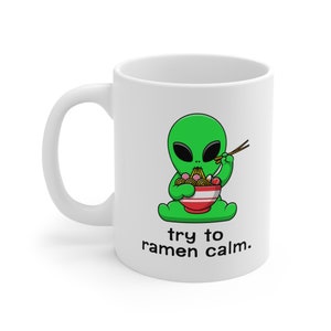 May include: White ceramic mug featuring a green alien cartoon character enjoying a bowl of ramen noodles with chopsticks. The text on the mug reads "try to ramen calm."