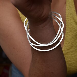 May include: A set of three silver-colored, irregularly shaped bangles. The bracelets are worn on a wrist, with the light reflecting off the metal. The background is blurred, with a yellow top and skin tones visible.