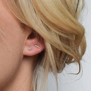 Könnte beinhalten: Eine Frau mit blonden Haaren trägt ein Paar goldene Ohrringe. Der eine Ohrring ist ein kleiner, dünner Stab und der andere ist ein winziger Diamant.