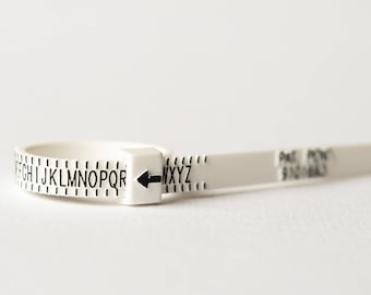 Pode incluir: Um medidor de tamanho de anel de plástico branco com marcas pretas e uma seta apontando para a direita. O medidor é marcado com letras de E a Z.