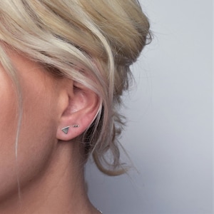 May include: A close-up of a person's ear with two small silver earrings. The earring on the lower lobe is a triangle shape, and the earring on the upper lobe is a small bar.