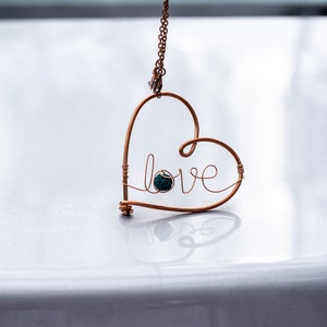 May include: Copper wire heart pendant necklace with the word "love" and a small, round, teal-colored stone. The heart-shaped pendant hangs from a delicate copper chain. The necklace is displayed on a reflective surface.