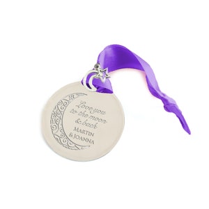 May include: A silver ornament with a crescent moon design and the text "Love you to the moon & back Martin & Joanna". The ornament is attached to a purple ribbon.