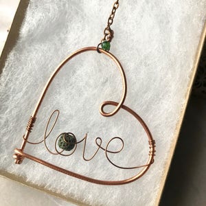 May include: Copper wire heart-shaped ornament with the word "love" spelled out in the wire. A small green and brown lava rock bead is in the centre of the heart.