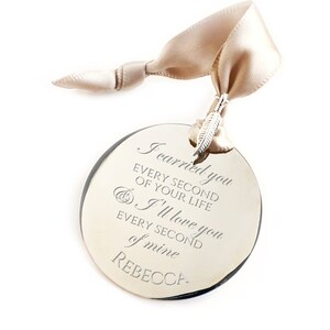 May include: A round, silver-coloured ornament with a beige ribbon. The ornament has the text "I carried you every second of your life I'll love you every second of mine REBECCA" engraved on it.