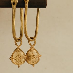 May include: A pair of gold-coloured earrings with a teardrop-shaped pendant. The pendant has a dotted pattern and small decorative elements. The earrings are suspended from a dark brown rod.