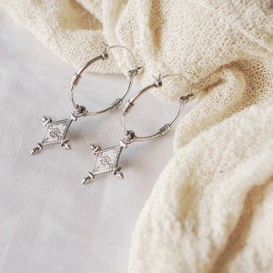 May include: A pair of silver hoop earrings with a diamond-shaped pendant. The pendant has an intricate, etched design and is suspended from the hoop. The earrings are displayed on a white, textured surface.