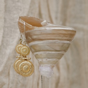 May include: Gold-toned earrings with a circular, sunburst design hang next to a seashell. The earrings have a layered, ornate appearance. The seashell is beige and white, with a spiral shape. The background is a soft, neutral fabric.