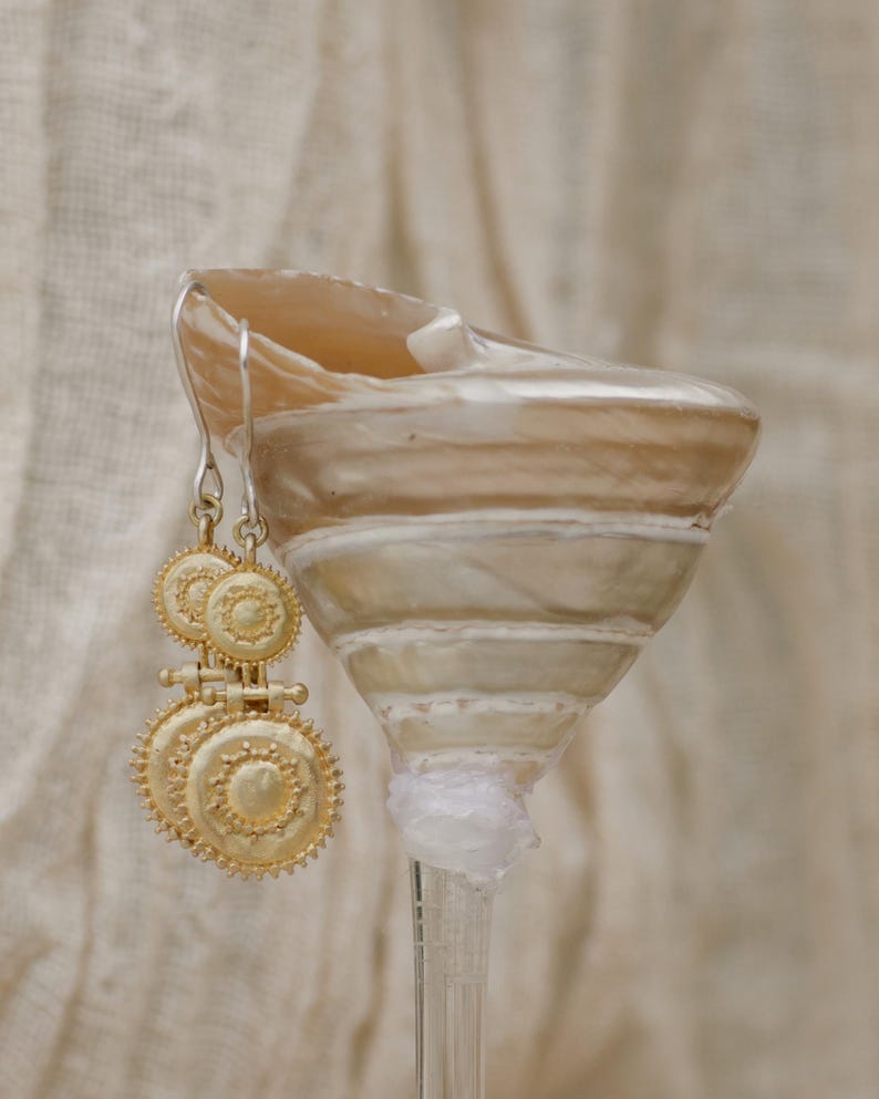 May include: Gold-toned earrings with a circular, sunburst design hang next to a seashell. The earrings have a layered, ornate appearance. The seashell is beige and white, with a spiral shape. The background is a soft, neutral fabric.