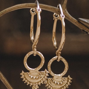 May include: A pair of gold-toned hoop earrings featuring a textured, irregular surface. Each hoop connects to a circular charm with a semi-circular, fan-shaped pendant attached at the bottom, decorated with small, raised spherical bead accents.