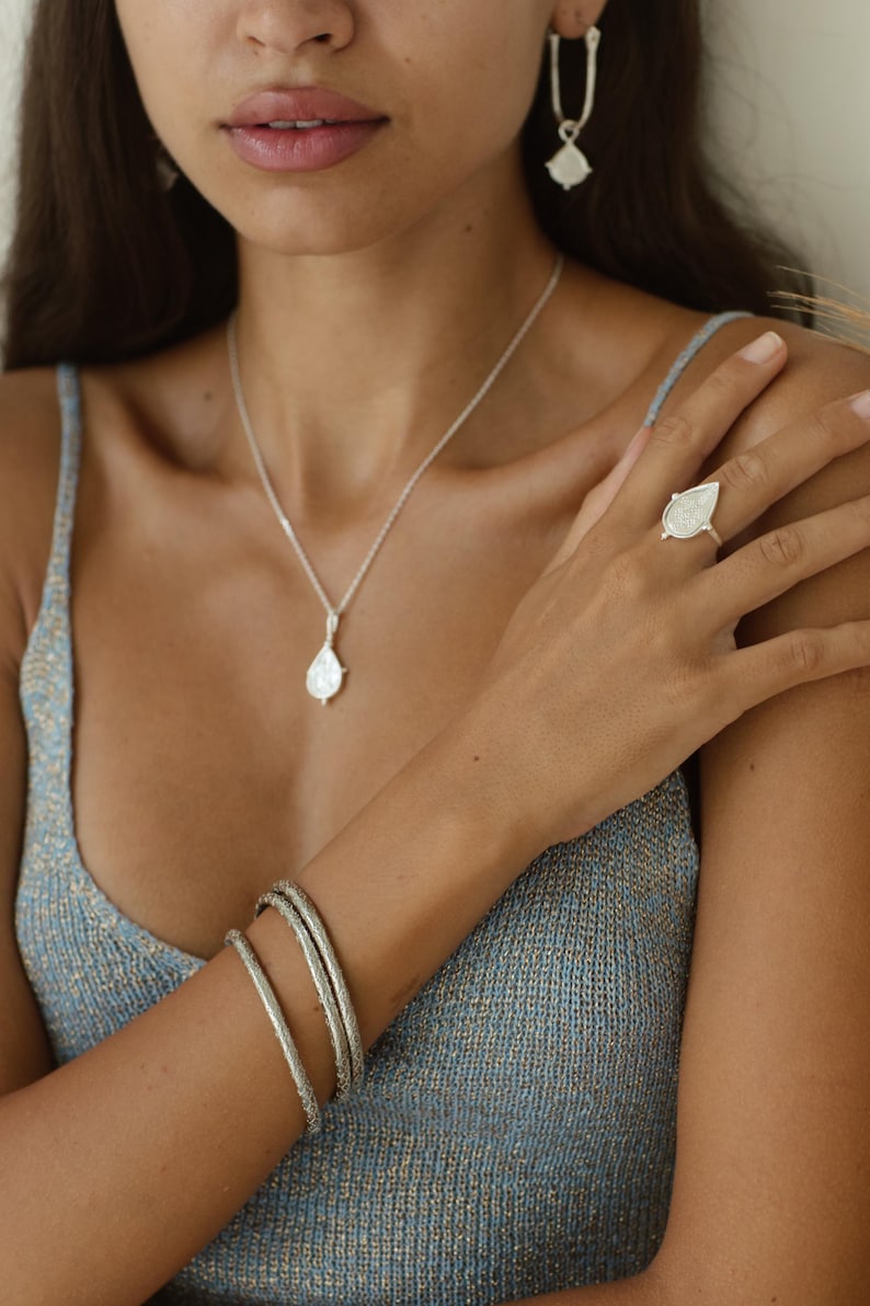 Puede incluir: Un collar de plata con un colgante en forma de l&aacute;grima, un anillo de plata con un dise&ntilde;o en forma de l&aacute;grima y un conjunto de tres pulseras de plata. Las joyas las lleva una persona con una blusa azul.