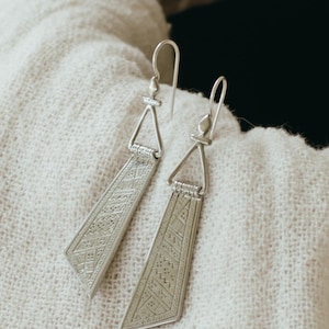 May include: A pair of silver dangle earrings with a geometric pattern. The earrings are made of a silver metal and have a triangular shape at the top. The earrings are hanging from a hook.