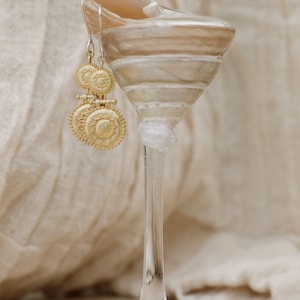 May include: Gold-toned earrings with a circular design, hanging from a clear glass. The earrings have a detailed, sunburst-like pattern. The glass is resting on a neutral-toned surface, with a soft, textured backdrop.