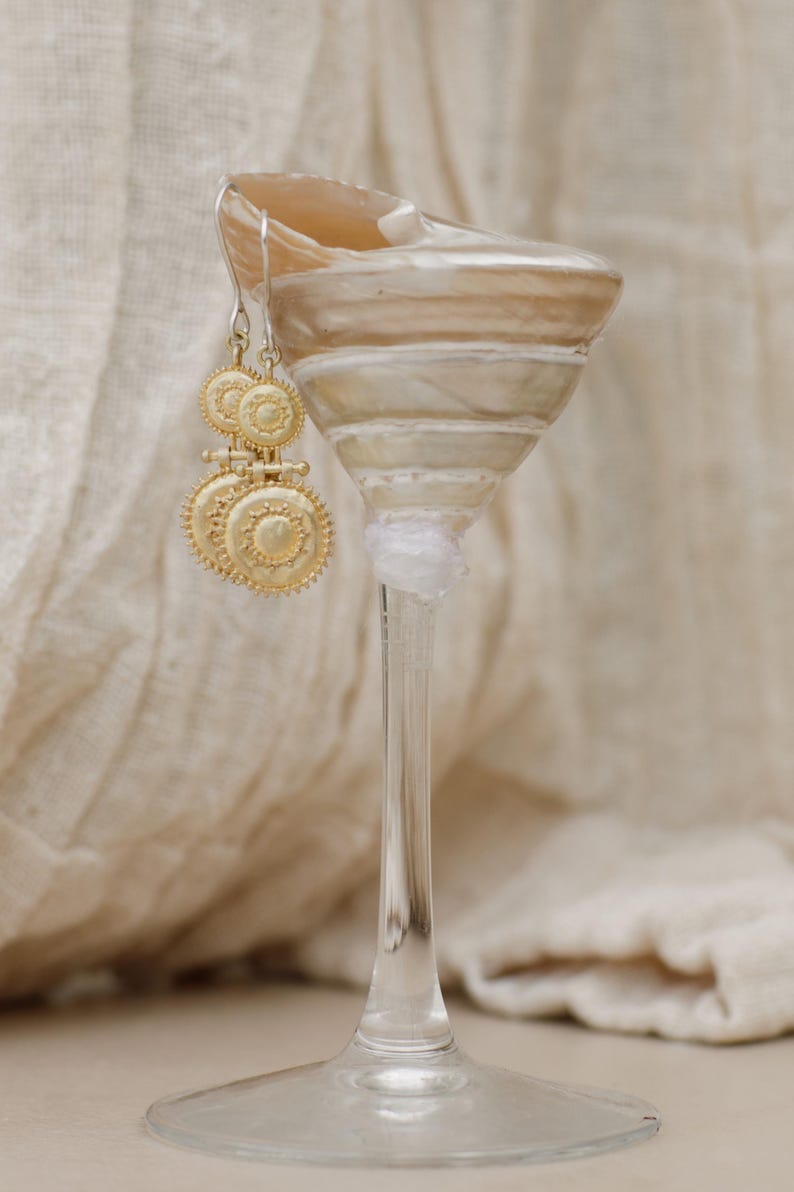 May include: Gold-toned earrings with a circular design, hanging from a clear glass. The earrings have a detailed, sunburst-like pattern. The glass is resting on a neutral-toned surface, with a soft, textured backdrop.
