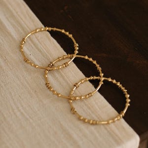 May include: Three gold bangles with a beaded design rest on a cream cloth. The bangles, made of metal, have a warm, golden hue. The background is a dark brown wooden surface. The bangles are likely to be approximately 6 cm in diameter.