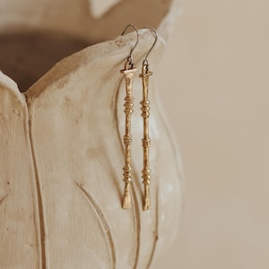 May include: A pair of long, slender, gold-toned metal drop earrings featuring a textured, rod-like design with small, decorative horizontal bands and rings along the length. The earrings hang from thin, curved metal ear wires.