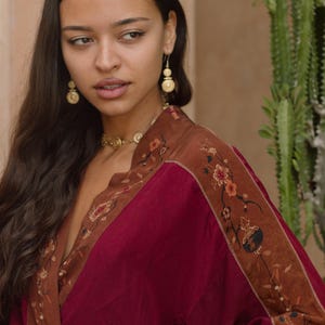 May include: A burgundy and brown kimono-style robe with floral embroidery. The robe has a deep V-neck and is paired with gold earrings and a matching necklace. The sleeves and edges are trimmed with a brown floral pattern.
