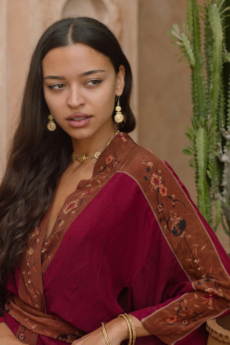 May include: A burgundy and brown kimono-style robe with floral embroidery. The robe has a deep V-neck and is paired with gold earrings and a matching necklace. The sleeves and edges are trimmed with a brown floral pattern.