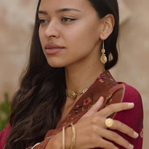 May include: A woman wearing gold jewelry, including a necklace, earrings, ring, and bracelets. The jewelry features circular designs. She is wearing a maroon top with a floral pattern. Her dark hair is long and wavy.