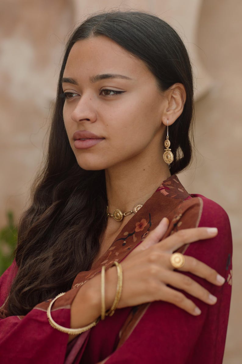 May include: A woman wearing gold jewelry, including a necklace, earrings, ring, and bracelets. The jewelry features circular designs. She is wearing a maroon top with a floral pattern. Her dark hair is long and wavy.