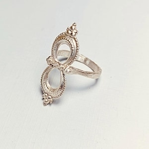 May include: A silver-colored ring with an openwork design. The ring features two oval-shaped elements connected by a band, each with a beaded border and a small floral-like embellishment at the top and bottom.