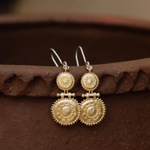 May include: A pair of gold-colored drop earrings. Each earring features a circular design with a smaller circle on top and a larger circle below, connected by a small bar. The earrings have a textured, antique appearance and are suspended from silver-colored hooks.