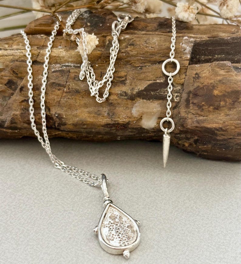 Puede incluir: Un collar de plata con un colgante en forma de l&aacute;grima, con una cadena delicada y un dise&ntilde;o texturizado. El colgante tiene un peque&ntilde;o bucle en la parte superior para sujetarlo a la cadena. El collar tambi&eacute;n incluye una cadena de plata con un peque&ntilde;o c&iacute;rculo y un extremo puntiagudo.