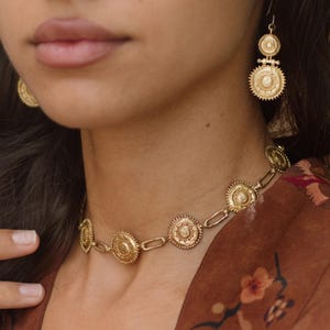May include: Gold-toned jewelry set featuring a necklace and earrings. The necklace has circular pendants linked by chain segments. The earrings have a tiered design with similar circular elements. The jewelry is worn against a brown floral patterned background.