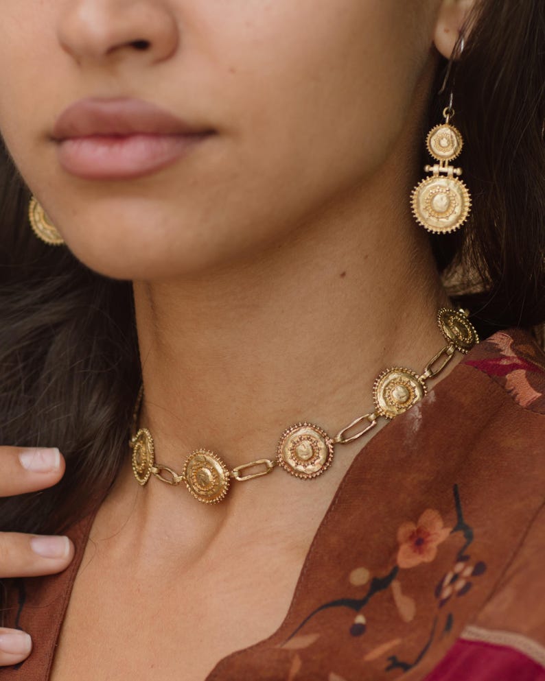 May include: Gold-toned jewelry set featuring a necklace and earrings. The necklace has circular pendants linked by chain segments. The earrings have a tiered design with similar circular elements. The jewelry is worn against a brown floral patterned background.