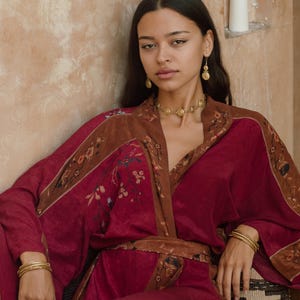 May include: A burgundy kimono-style robe with brown floral trim and a matching belt. The robe is paired with gold jewelry, including a necklace, earrings, bracelets, and a ring. The overall aesthetic is bohemian and elegant.
