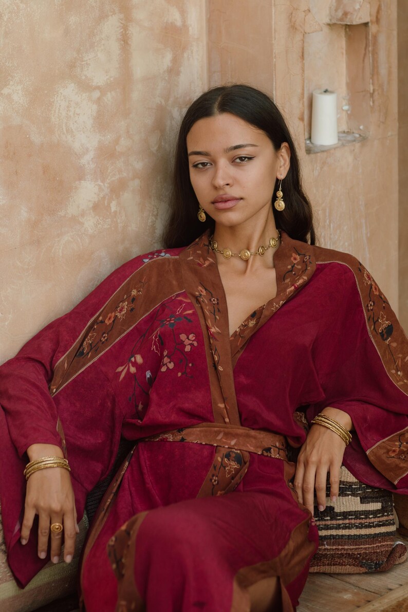 May include: A burgundy kimono-style robe with brown floral trim and a matching belt. The robe is paired with gold jewelry, including a necklace, earrings, bracelets, and a ring. The overall aesthetic is bohemian and elegant.
