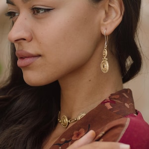 May include: A woman wearing gold-colored jewelry, including earrings, a necklace, and a ring. The earrings are long and dangle, featuring a circular design. The necklace and ring also have a similar circular design. The woman is wearing a maroon floral patterned top.