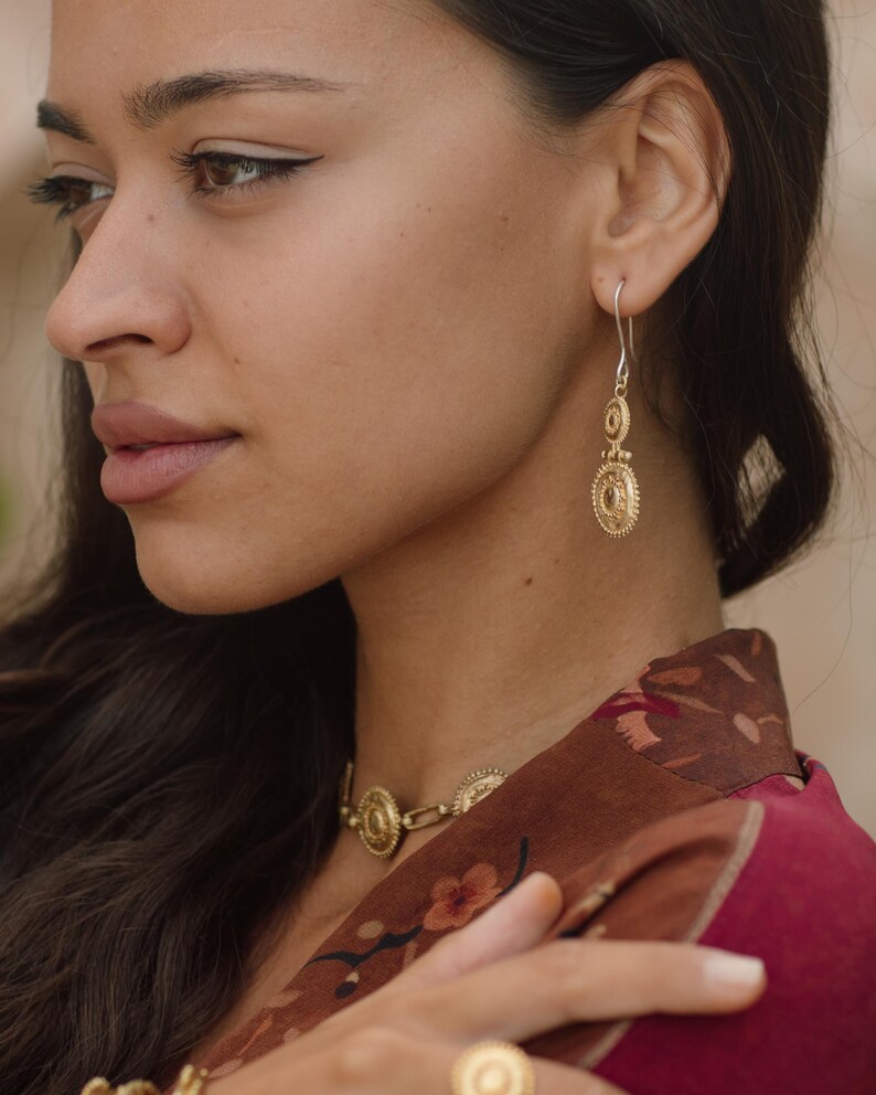 May include: A woman wearing gold-colored jewelry, including earrings, a necklace, and a ring. The earrings are long and dangle, featuring a circular design. The necklace and ring also have a similar circular design. The woman is wearing a maroon floral patterned top.