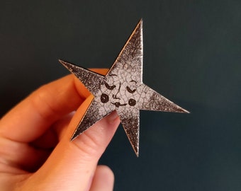 May include: A silver star-shaped ornament with a black and white face drawn on it. The star has a textured surface.