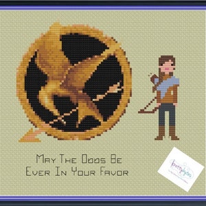 Pode incluir: Padrão de ponto cruz com um alfinete dourado de mockingjay com um design de pássaro estilizado e as palavras "Que a sorte esteja sempre ao seu lado". Uma personagem feminina com cabelo castanho e um arco e flecha está à direita do alfinete.