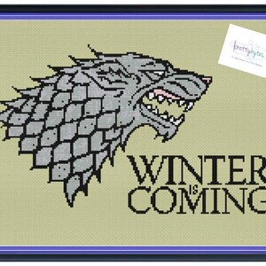 May include: Cross-stitch artwork featuring a gray direwolf head with open jaws, set against a light green background. The text "WINTER IS COMING" is below the wolf. The piece is framed in black with a purple inner border.