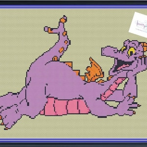 Pode incluir: Ponto cruz de um dragão roxo com detalhes laranja, emoldurado em preto e azul. O dragão está deitado, sorrindo. O fundo é bege claro.