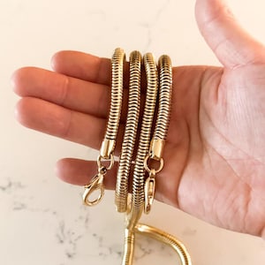 Puede incluir: Una cadena dorada y flexible con un lazo y dos cierres. La cadena está hecha de una serie de segmentos acanalados conectados. La cadena se sostiene en una mano sobre un fondo de mármol blanco.