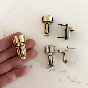 May include: A selection of metal purse clasps in gold and silver finishes are shown on a white marble surface. The clasps have a rectangular design with a curved opening. Some are two-part, others are single pieces.
