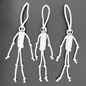 May include: Three handmade white skeleton ornaments. Each skeleton has a round head with a painted skull face, a woven body, and dangling arms and legs. The ornaments are suspended by a loop of white cord.