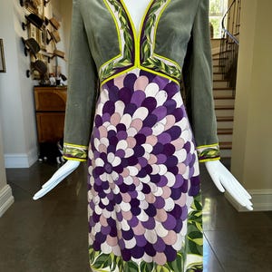 May include: A vintage-style dress with a green velvet bodice and long sleeves. The dress features a V-neck with leaf-patterned trim and a skirt with a floral design in shades of purple, white, and green. The dress is displayed on a mannequin.