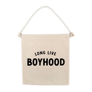 May include: A beige canvas banner with a wooden dowel at the top and a rope loop for hanging. The banner has black text that reads "Long Live Boyhood".