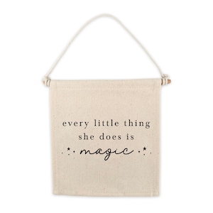 May include: A beige canvas wall hanging with the text "every little thing she does is magic" in black lettering with small stars on either side of the word "magic".